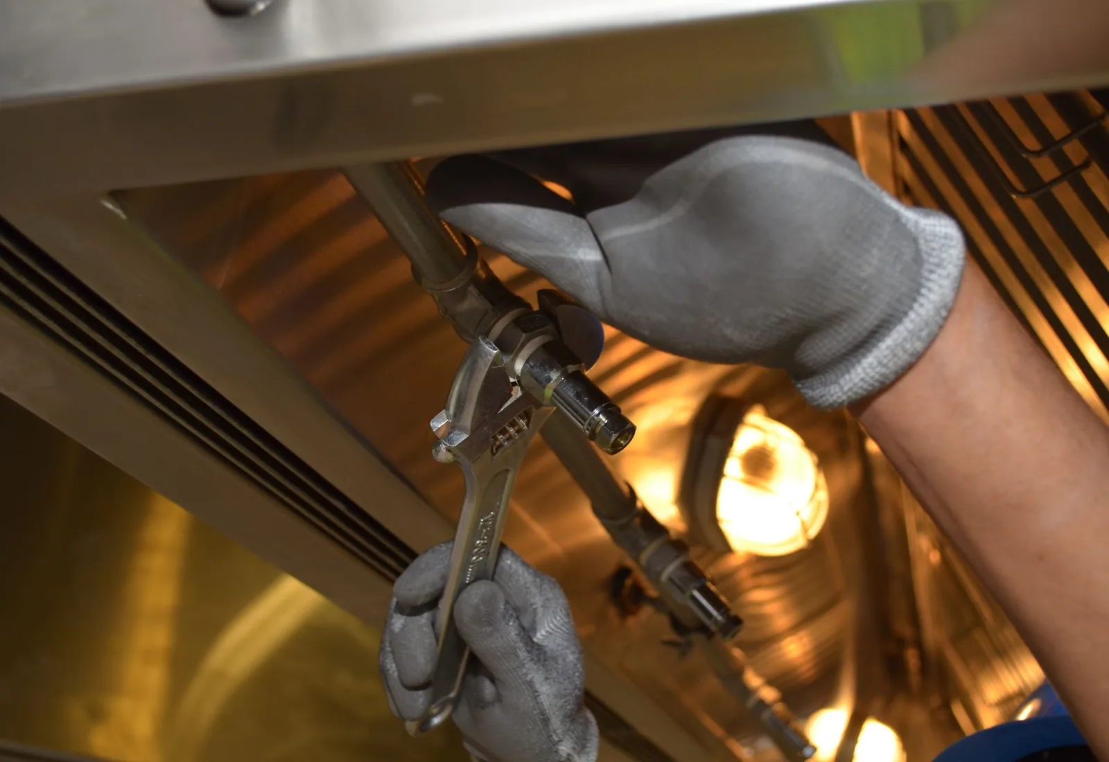 A technician in work gloves tightening a fitting on a water heater supply line with an adjustable wrench